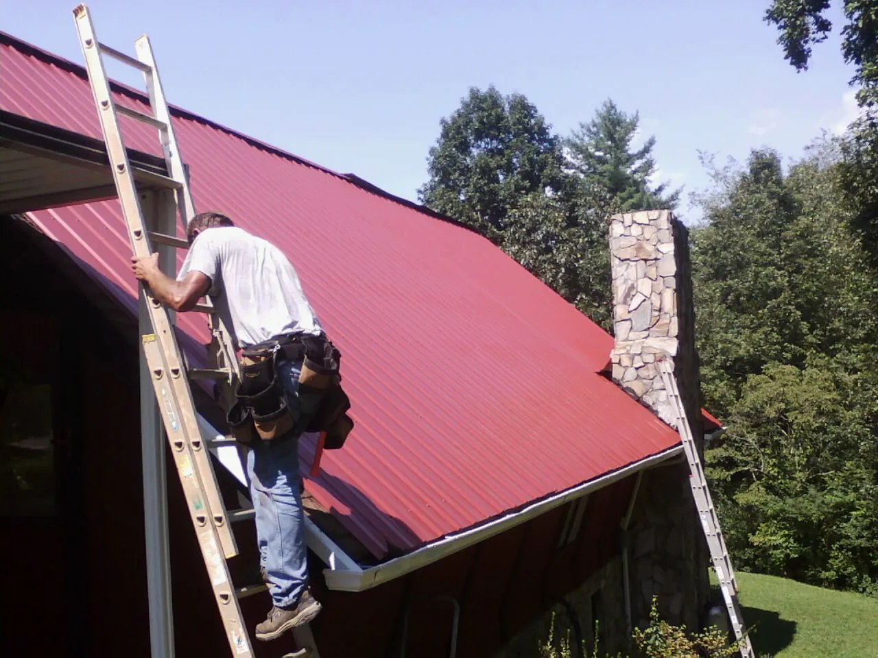 Roofer on a ladder beside a red metal roof and stone chimney — Soffit & Fascia Repair by Roofing Brothers in Hayward